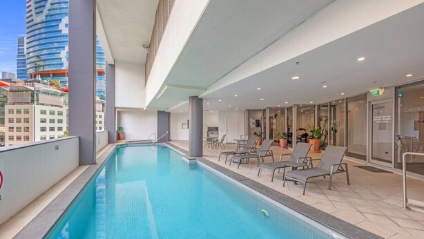A modern indoor pool with clear blue water, surrounded by lounge chairs and a glass wall offering a city view. - Dreamy Cityside 2BR Haven Brisbane Central (Brisbane City)