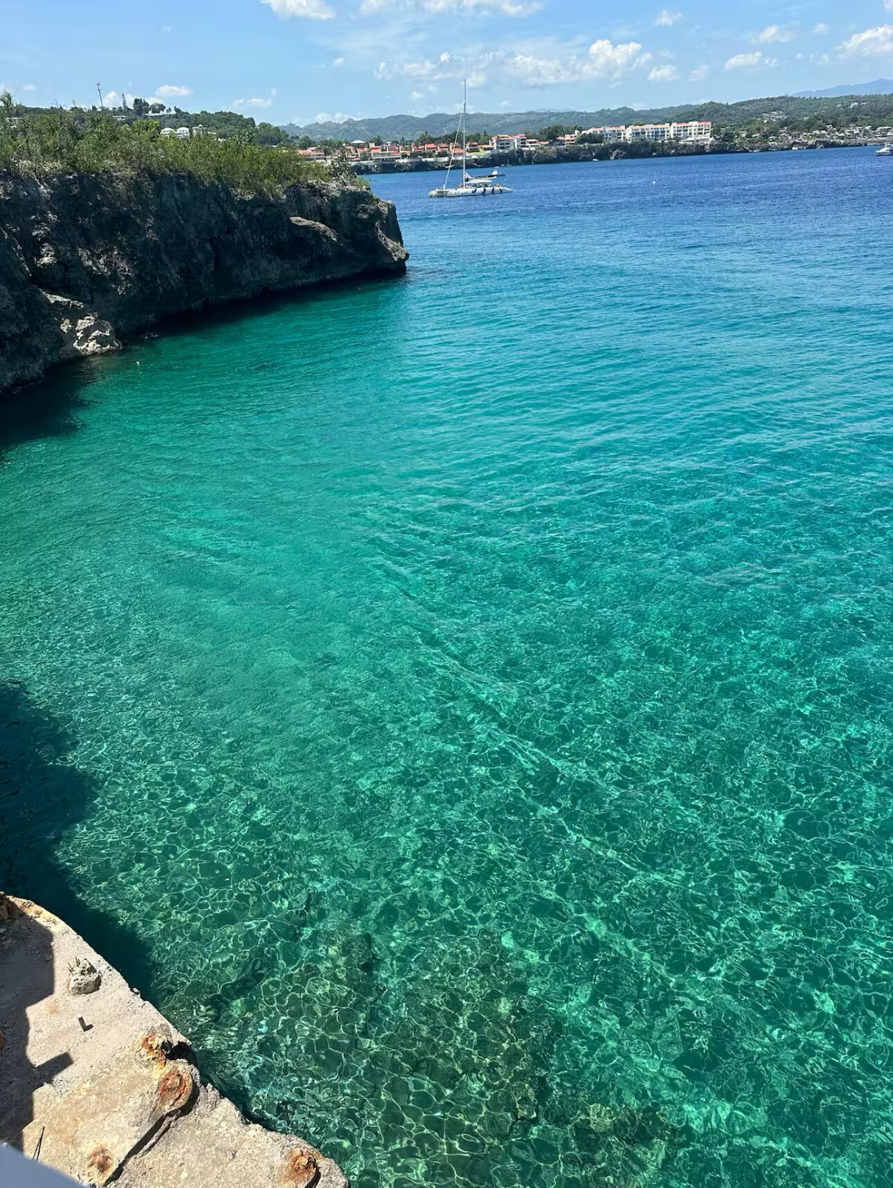 En kustvy med klart turkosfärgat vatten, en stenig strand och en byggnadskomplex i fjärran.