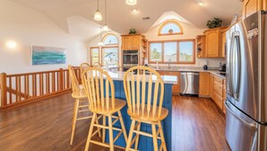 A kitchen with wooden cabinets, a stainless steel refrigerator, and a bar area with wooden chairs. - Seventh Heaven - Corolla, Ocean Sands, 500 Yds to Beach, Pool, Hot Tub, Pool Table, Pets OK (Corolla)