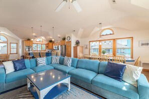 Ein geräumiges Wohnzimmer mit einem Ecksofa, einem Essbereich mit Holzsesseln, einer Küche mit Holzschränken und einem Deckenventilator.