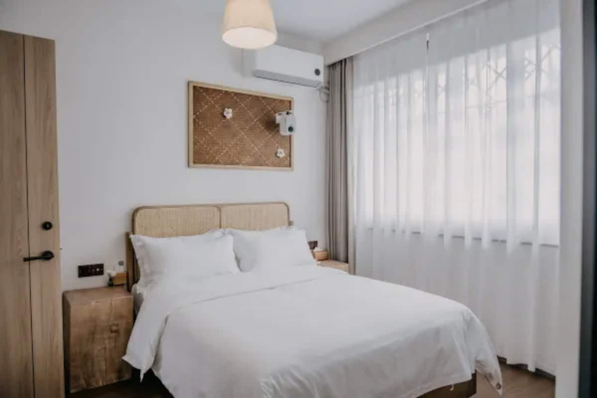 A bedroom with a bed, wooden nightstand, wall-mounted air conditioning unit, and a window with sheer curtains.