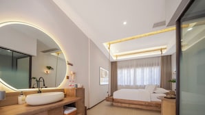 Quarto de hotel moderno com uma cama grande, uma mesinha de cabeceira de madeira com um espelho amplo e uma janela grande com cortinas.