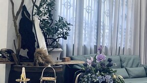 Uma sala de estar com um sofá verde, uma mesa de centro com um arranjo floral e uma janela com cortinas leves.