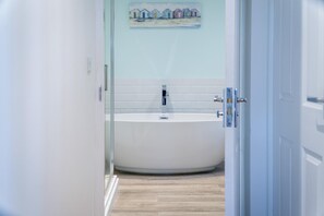 A modern bathroom with a freestanding bathtub, white tiled walls, and a wooden floor. - Caldey View Cottage - 1 Bedroom - Penally (Tenby)