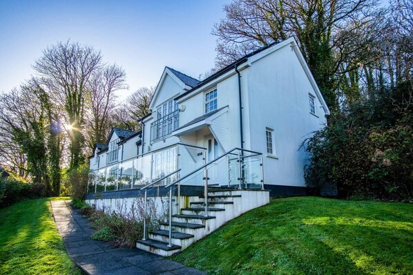 A modern two-story house with a glass staircase leading to the entrance, surrounded by lush greenery and trees. - Caldey View Cottage - 1 Bedroom - Penally (Tenby)