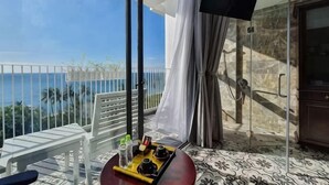 Een balkon met uitzicht op de oceaan, een witte stoel, een dienblad met een drankje, en een glazen deur naar een doucheruimte.