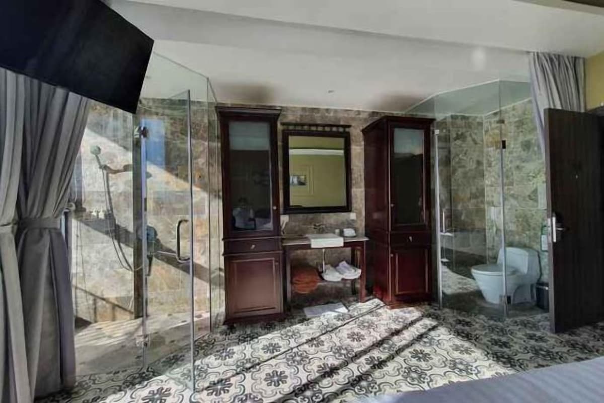 Hotelkamer met een glazen douchecabine, een houten wastafelmeubel met spiegel, een bed met een beddengoed met een patroon, en een badkamer met toilet en wastafel.