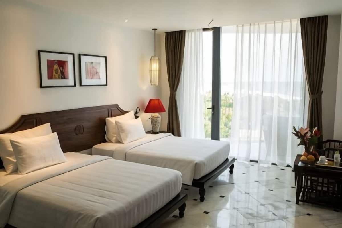 Ein Hotelzimmer mit zwei Betten, einem Nachttisch mit Lampe, einem Essbereich mit Tisch und Stühlen sowie einem großen Fenster mit Vorhängen.