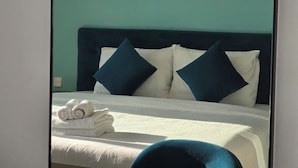 A neatly made bed with white linens and two dark blue pillows, a folded white towel on the bed, and a teal-colored wall in the background. - Marina Hillside Condotel (An Thoi)