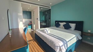 A modern bedroom with a large bed, a wooden table, and a kitchenette in the background. - Marina Hillside Condotel (An Thoi)
