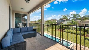 Ein Balkon mit einer dunklen Rattan-Couch, einem Pool und Blick auf angrenzende Häuser und Grünflächen.