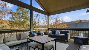 Un espacio de estar al aire libre cubierto con una mesa y sofás, con vistas a un paisaje de árboles y un edificio lejano.