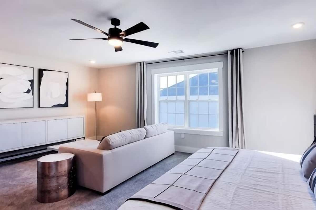 A modern bedroom with a bed, a sofa, a standing lamp, a ceiling fan, and a window with curtains.