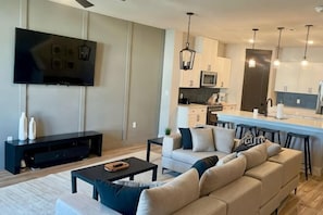 A modern living room with a sofa, coffee table, and TV. A kitchen with a microwave and bar stools is visible. - Loop, New Luxurious Large 3BR, Sleeps 9 + Parking (Collin County)