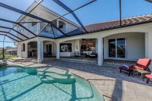 A covered outdoor swimming pool area with a patio, lounge chairs, and a house in the background. - Villa Skye (Cape Coral)