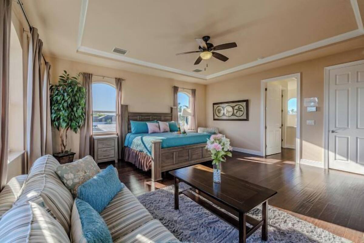 A bedroom with a bed, a sofa, a coffee table, a ceiling fan, and a large window.