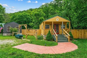 A small wooden cabin with a porch and steps, surrounded by a wooden fence and lush greenery.