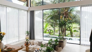 A modern living room with a large window overlooking a tropical garden and a pool. - Dzen Greenfield Villa 7 (Pejeng)