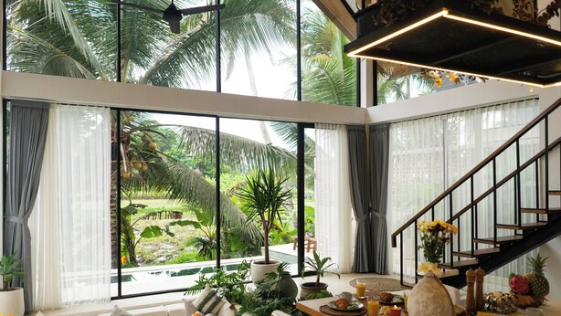 A spacious living area with a large window overlooking a lush garden, a wooden dining table with breakfast items, and a staircase leading to an upper level. - Dzen Greenfield Villa 7 (Pejeng)