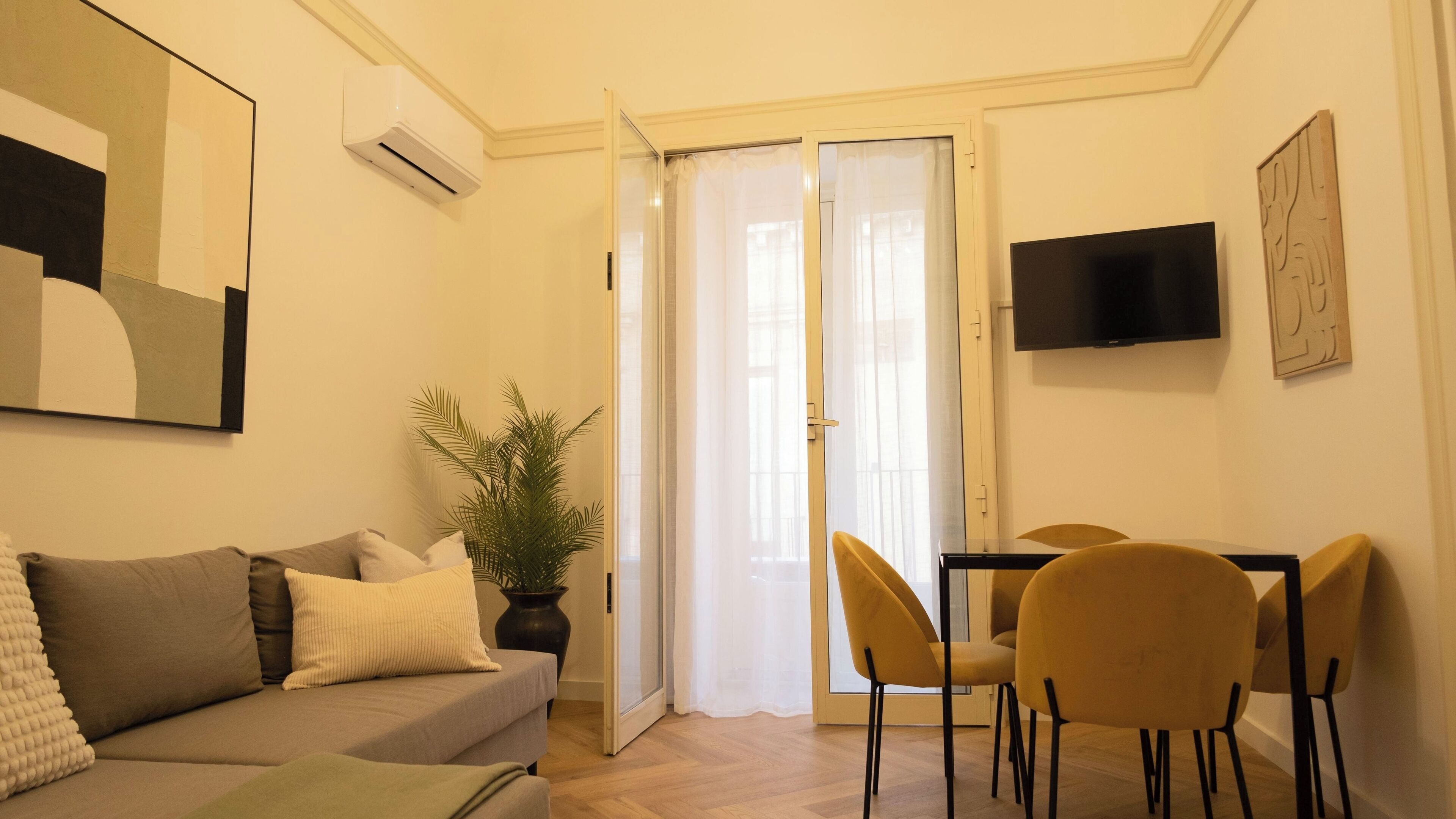 Una sala de estar con un sofá, una mesa de comedor con sillas, un televisor montado en la pared y una planta en maceta.
