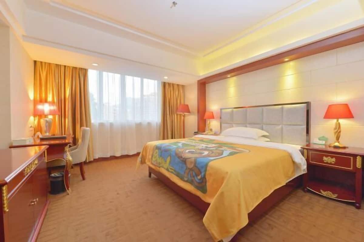 A hotel room with a large bed, two bedside lamps, a desk with a chair, and a view of the city through large windows.