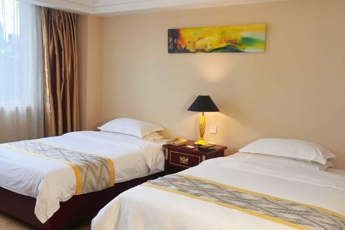 A hotel room with two beds, a painting on the wall, a lamp on a nightstand, and a window with curtains.