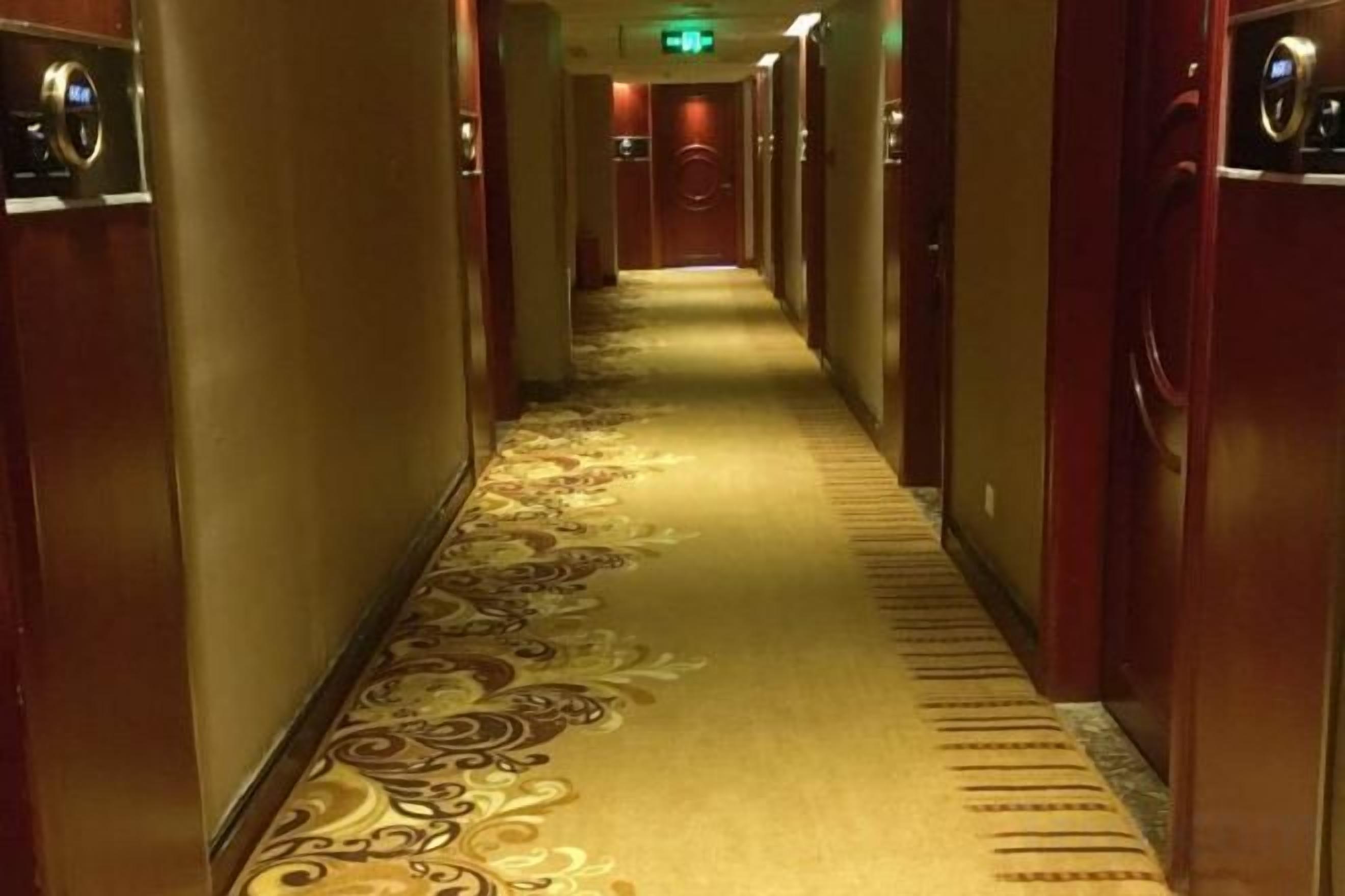 A hotel hallway with carpeted floors, doors on both sides, and a green exit sign at the end.