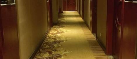 A hotel hallway with carpeted floors, doors on both sides, and a green exit sign at the end.