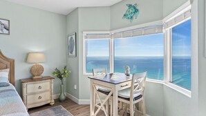 A bedroom with a bed, bedside table, lamp, framed artwork, a vase with flowers, a dining table with chairs, and a large window with a view of the ocean. - Gulf-front PCB Condo w/ Balcony, Resort Amenities, Game Room, Pools & Beach Gear Tortuga 1301 by Avantstay (Panama City Beach)