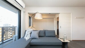 Une chambre d’hôtel moderne avec un canapé gris, une petite table de chevet sur laquelle se trouvent une bouteille et un verre, et une grande fenêtre donnant sur des immeubles.