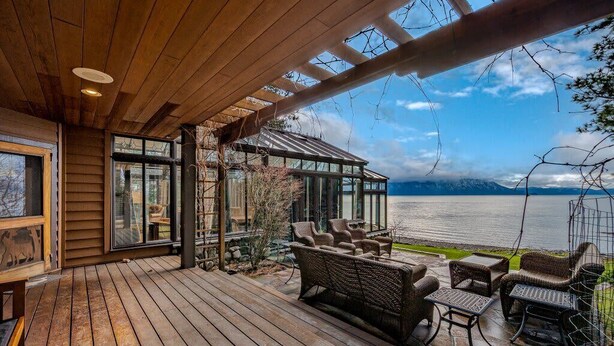 Un terrazzo in legno con mobili in vimini che si affaccia su un lago e montagne.