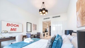 Una habitación de hotel moderna con una cama, un escritorio, una silla, una lámpara de escritorio, un espejo, una obra de arte en la pared, un letrero que dice 'BOOK ME', una zona de cocina y una puerta que da a otra habitación.