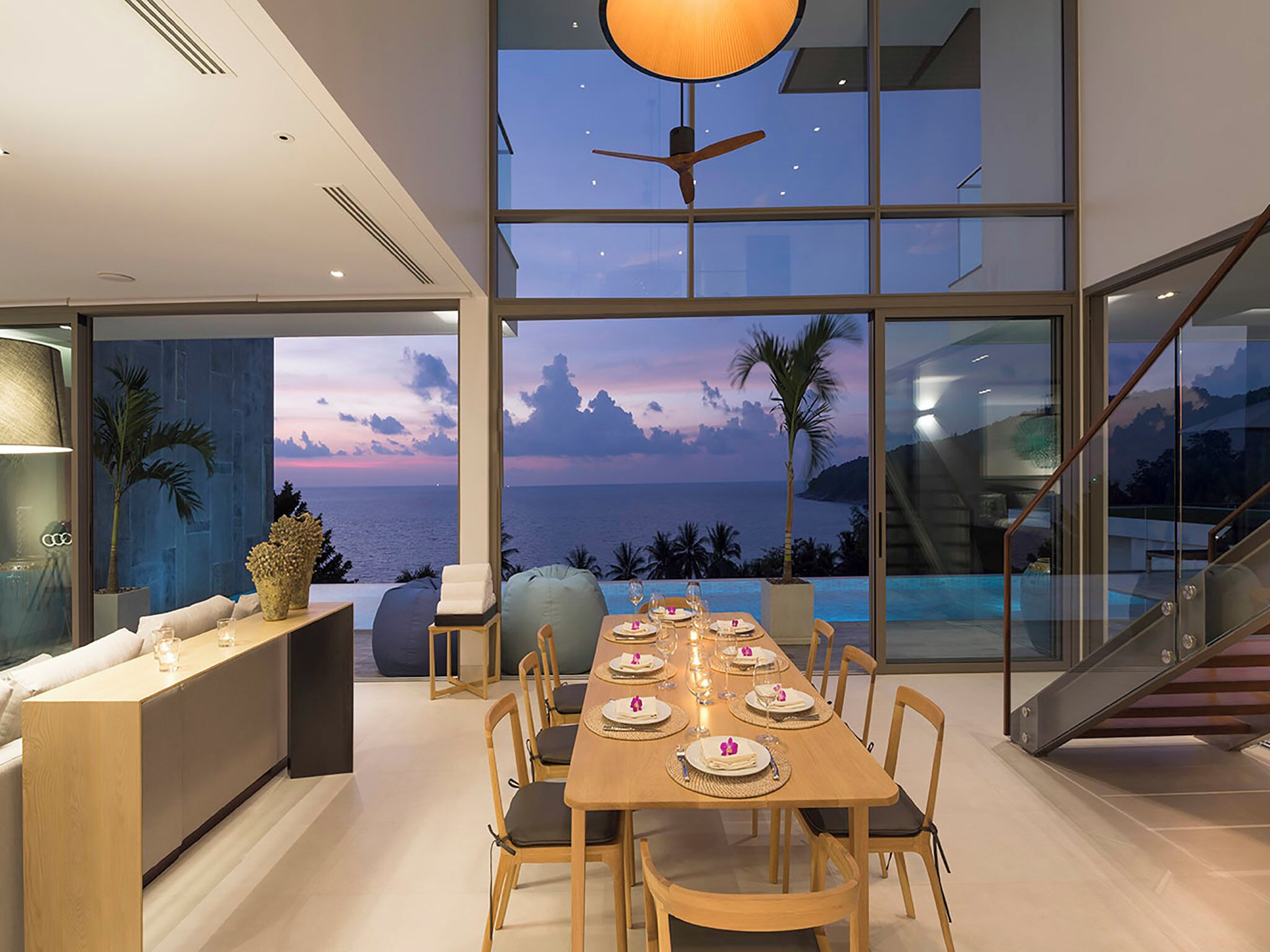 A modern dining area with a view of the ocean at sunset, a large window, and a ceiling fan.