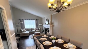 Une salle à manger avec une longue table dressée pour six personnes, un lustre au-dessus, et un salon à l’arrière-plan.