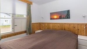 A bedroom with a large bed, wooden paneling on the walls, a window with curtains, a painting of a sunset, and a white dresser.