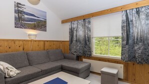 A modern living room with a grey sectional sofa, a white coffee table, and a small ottoman. There is a large window with forest-themed curtains, a wall-mounted lamp, and a framed landscape painting.