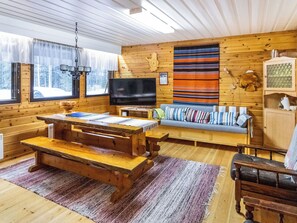 Interior acolhedor de uma cabana com uma mesa de jantar de madeira, um sofá com almofadas coloridas, uma televisão e uma janela grande com vista para uma paisagem coberta de neve.