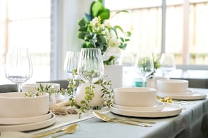 A dining table set with white plates, bowls, and clear glassware, adorned with greenery and a vase of flowers.