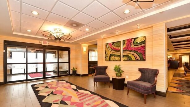 Un hall de hotel con una alfombra estampada, dos sillones, una mesita con una planta y un cuadro abstracto grande en la pared.