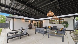 Un patio exterior con mobiliario moderno, incluyendo sillones y sofás, bajo una zona cubierta con luces colgantes.