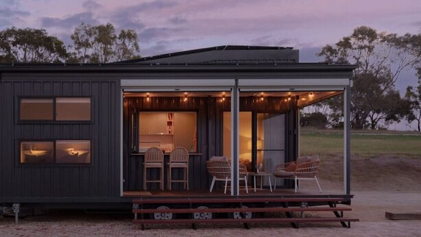 A modern cabin with a covered outdoor seating area, a fire pit, and a view of the surrounding landscape.
