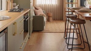 A modern kitchen with wooden cabinets, a built-in oven, a dining area with a bar stool, and a ladder leading to a loft bedroom.