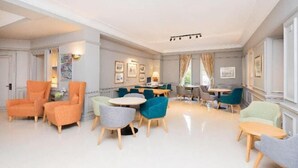 Un vestíbulo de hotel con varias zonas de estar, que incluyen mesas redondas y sillas, y un amplio ventanal que proporciona luz natural.