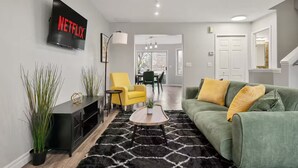 Una sala de estar moderna con un sofá verde, un sillón amarillo y un tapete blanco y negro. Un televisor de pantalla plana está montado en la pared, y se observa un comedor al fondo.