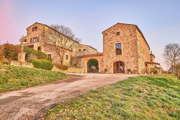A stone building with a central archway and a red door, surrounded by a gravel driveway and grassy areas.