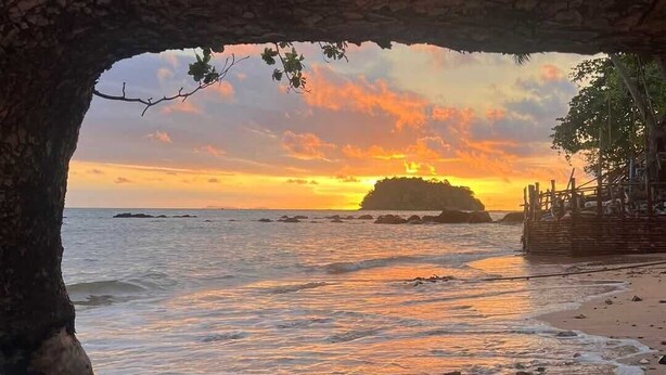 A beach scene with a natural rock arch framing the view of the ocean and a distant island at sunset. - Ban Slow Life Homestay Koh Libong (Ko Libong)