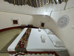A single bed with a colorful blanket, two pillows, and a rolled towel. There are two small wooden chairs and a small table. The room has a simple design with a patterned wall and a woven ceiling.