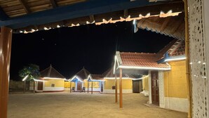 A courtyard with several buildings, each with a tiled roof and a door. The buildings are illuminated by lights, and there is a tree with colorful lights in the foreground. - Rann Sanji Stay (Bhuj)