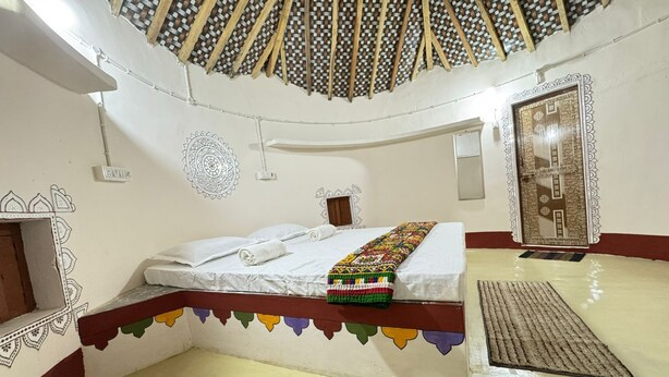 A room with a bed, a decorative wall, a door, and a ceiling with a woven pattern. - Rann Sanji Stay (Bhuj)