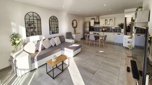 Ein modernes Wohnzimmer mit einem grauen Sofa, einem kleinen Holztisch und einem Essbereich mit Tisch und Stühlen. Im Hintergrund ist eine Küche mit weißen Schränken und einer grauen Arbeitsplatte erkennbar.